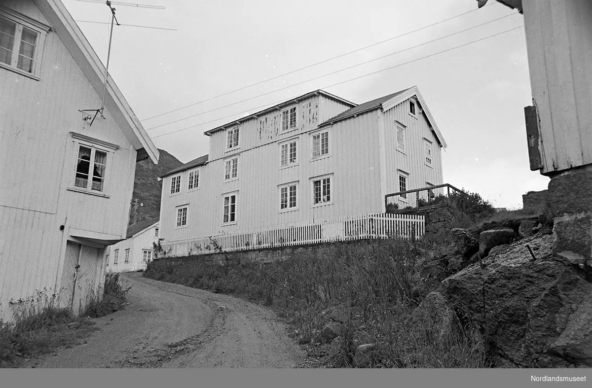 Bygninger i fiskeværet Nusfjord. Større bolighus på oppmurt steinmur. Vei foran. Hagegjerde langs kanten på steinmuren. Bygningen med mange smårutede vinduer oppe og nede. Større ark med 2 vinduer. Stående panel.