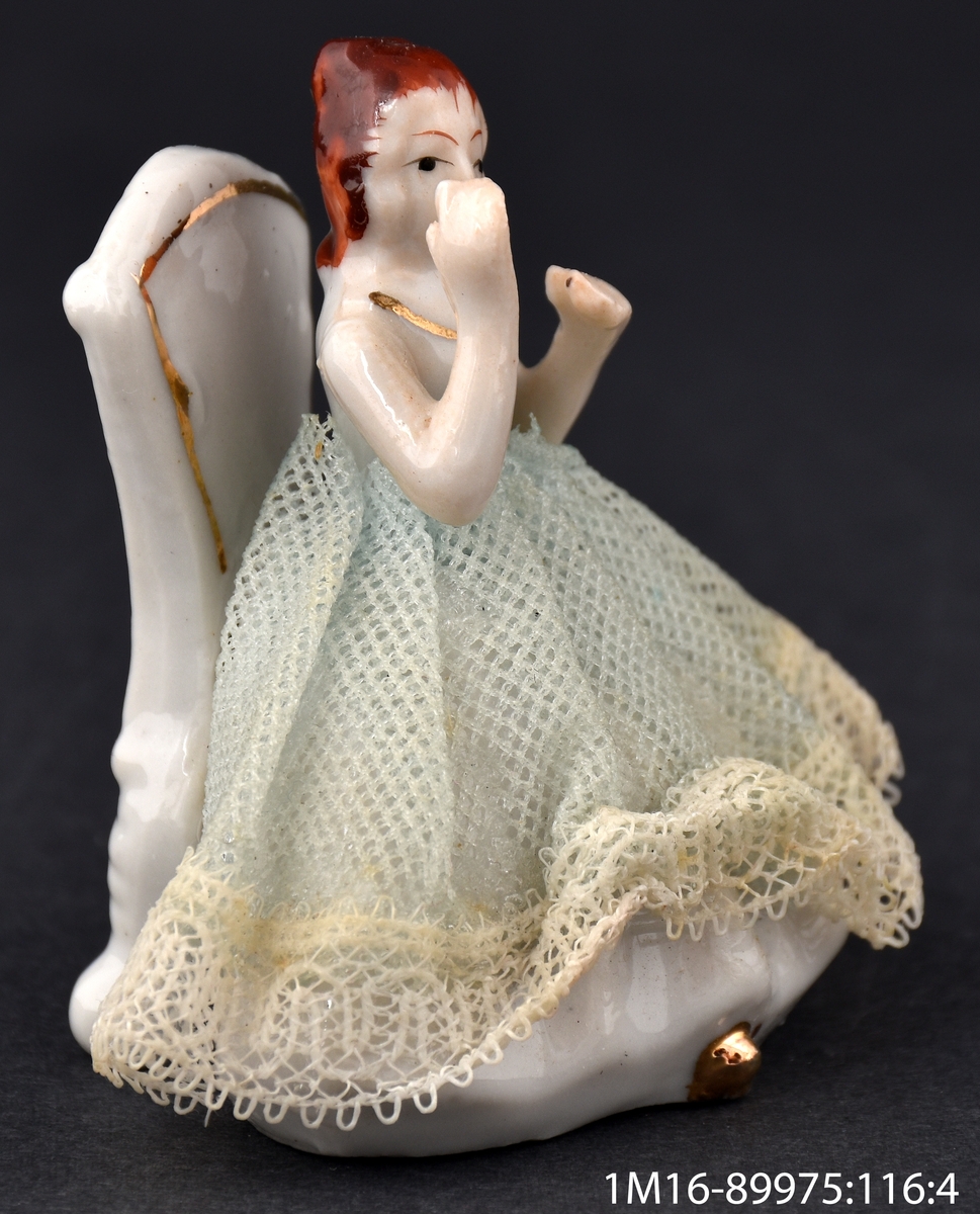 Leksak av porslin, i form av en dam som sitter på en stol i vitt och guld och applåderar. Damens kjol är gjord av veckat tyg med en spetskant nedtill.