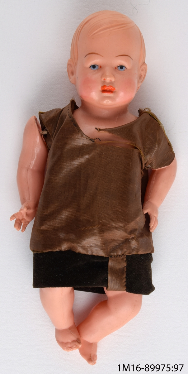 Babydocka av celluloid med rörliga armar och ben, klädd i brun klänning av vaxat siden med kant av sammet nedtill. Handsytt.