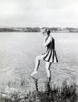 Svart-hvitt foto av ung kvinne iført badedrakt med skjørt, som putter tåa i vannet. (Foto/Photo)