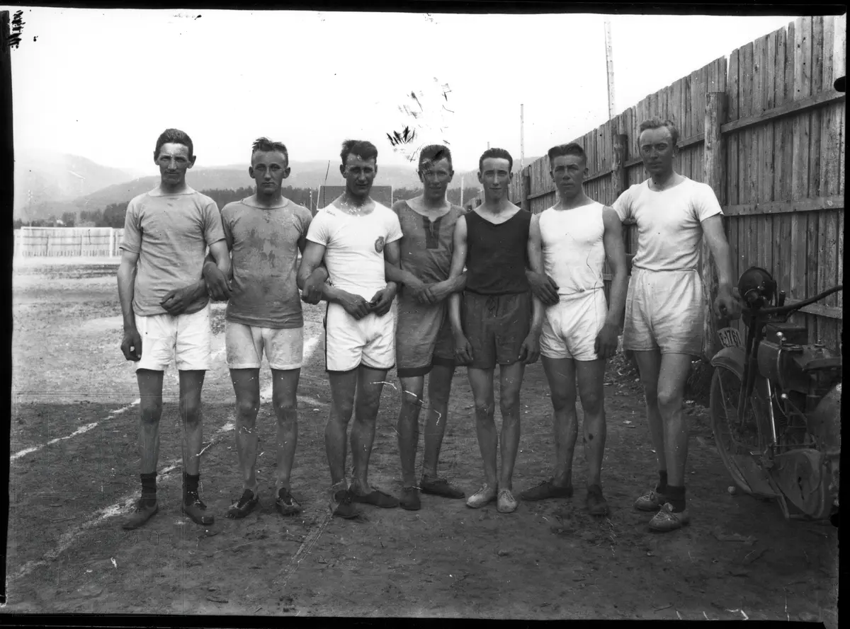 7 menn i kortbukse og trøye står samlet på en idrettsplass. JIF banen på Jevnaker.