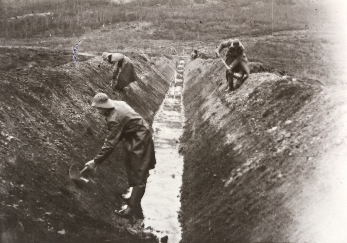 Tre menn i regnfraker arbeider på en vanningskanal. Kanalen i senter av bildet, 2 menn arbeider på venstre siden og 1 mann arbeider på høyre side. Skogkledd åsside.
