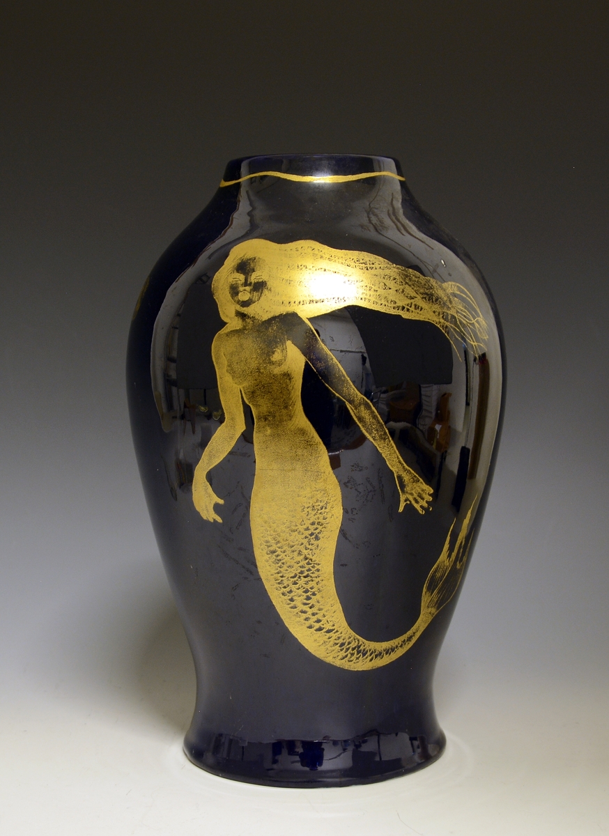 Prot: Stor vase av porselen. Dyp blå glasur. Dekor i gull: havfrue og fisk. Uten mrk. og sign. Form: Konrad Galaaen, dekor: Andor Hubay. Produsert 1954.