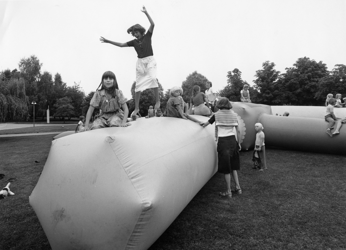 Studsmatta i Folkets Park i Linköping 1970-80-tal. Studsmatta i form av smala armar. Många barn sitter runt om på mattan, ett hoppar. I bakgrunden ser man lövträd.