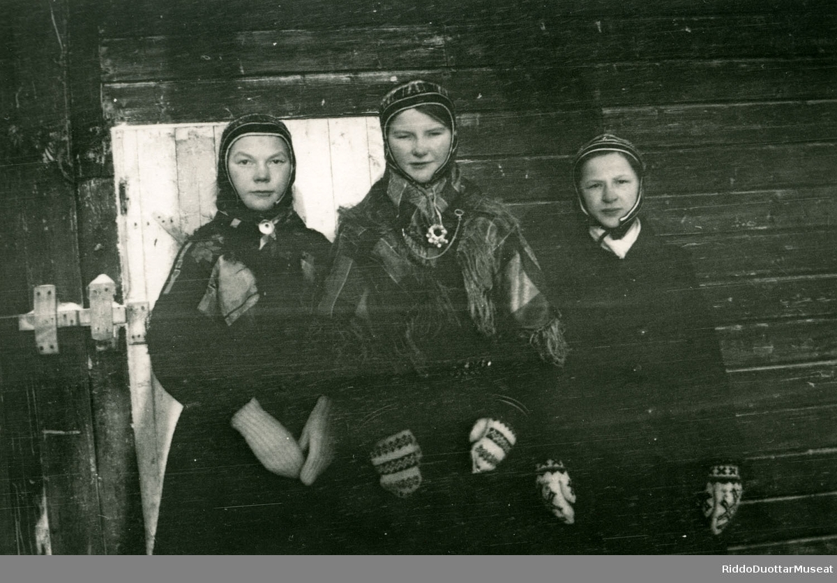Golbma sámi nieidda olggobealde viesu.
Tre jenter står ved veggen.