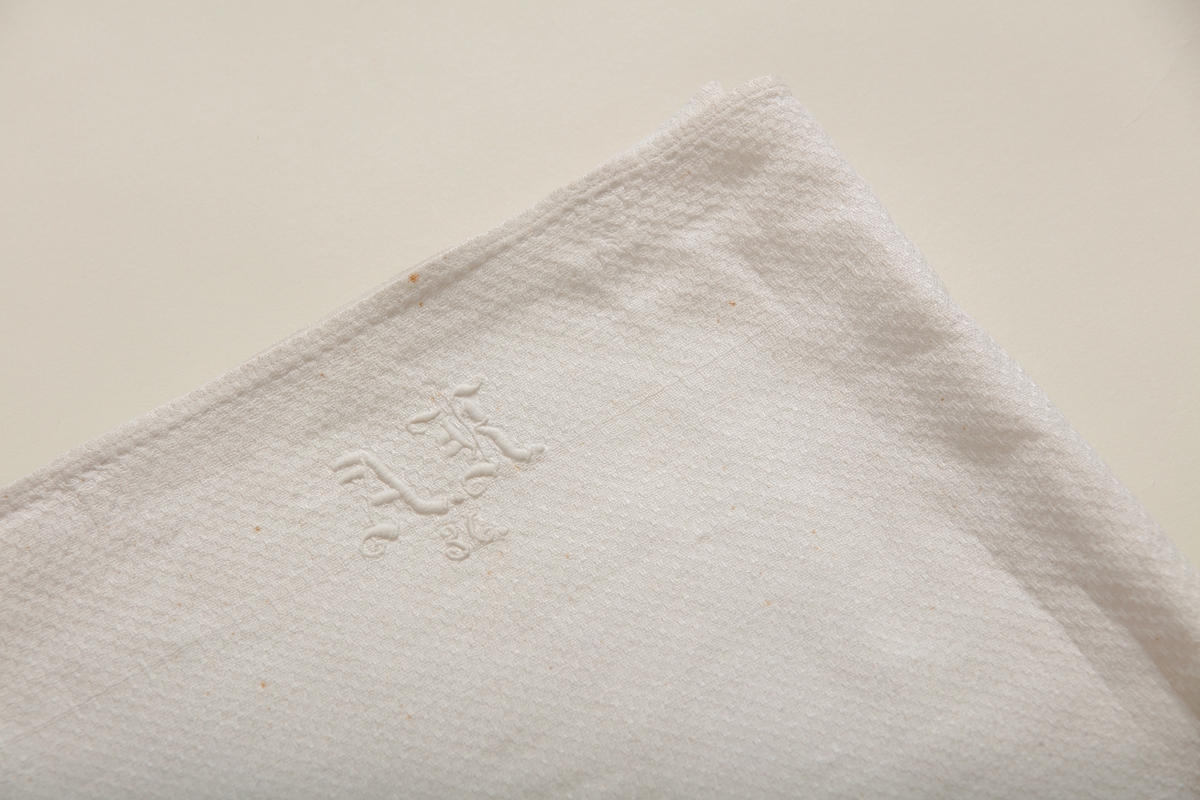 Håndkle av hvit lin med mønstereffekt i vevningen. Maskinsydd fold i hver ende. Hvitt håndbrodert monogram i plattsøm. Påsydd hank av bomullsbånd.