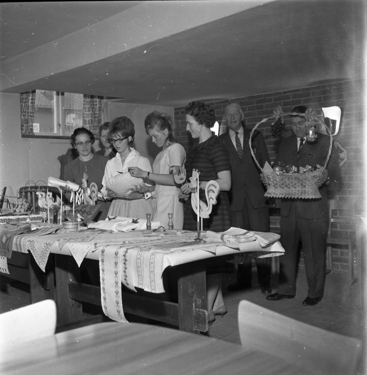 Fem kvinnor står bakom ett bord där det ligger flera broderade dukar. En man står och håller i en fruktkorg. Möjligen ett vinstbord till lotteri.
