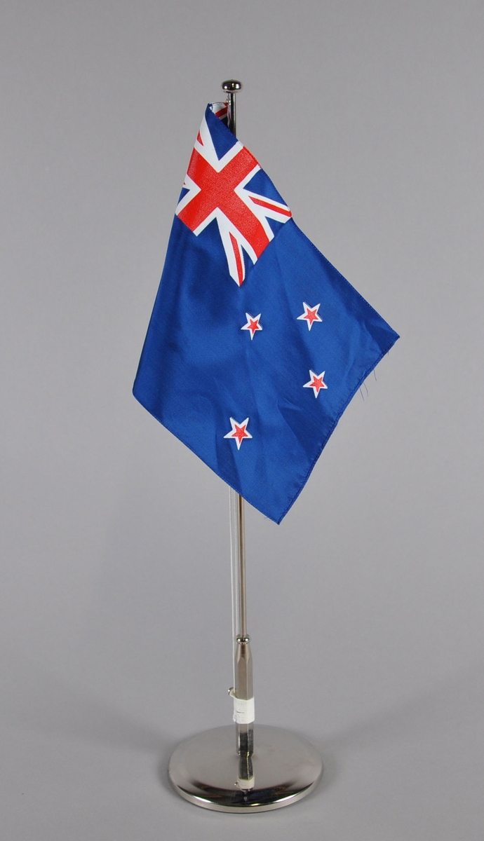 Bordflagg på stang fra New Zeland. Storbritannias flagg i øverste hjørne mot stangen, og fire hvite og røde stjerner på mørkeblå bakgrunn.