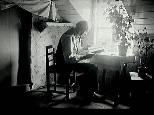 En gammal man sitter på en liten stol vid ett fönster och läser. På borden står pelargoner. Det är Åke Lundqvist, mannen som 1895 sålde sin gård till Skansen - Oktorpsgården.