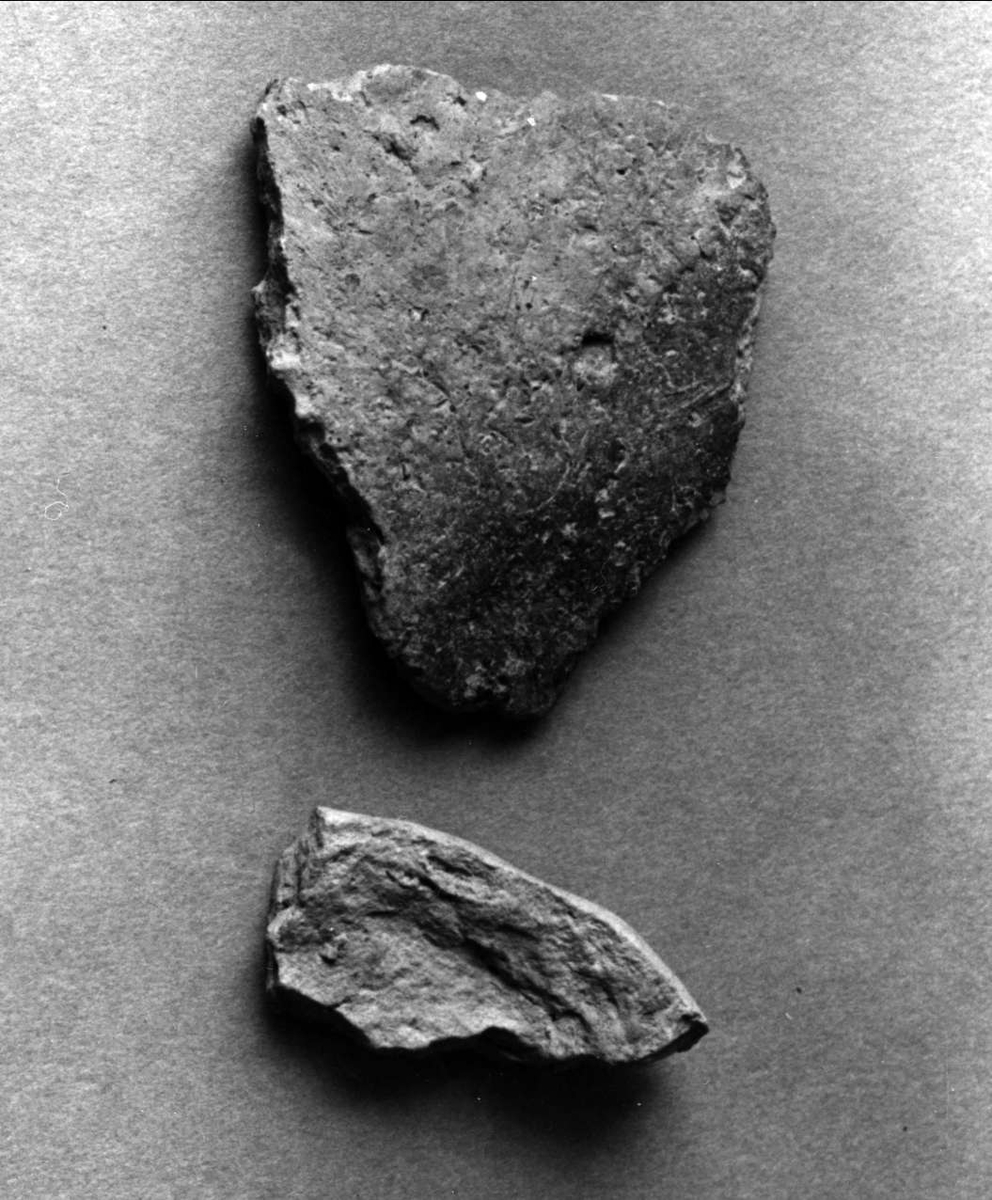 Fragment av kärl. Det större fragmentet är så kallat A-gods, med grovt magrad keramik. Det mindre fragmentet är bränd, formad lera, möjligen ett bottenfragment.