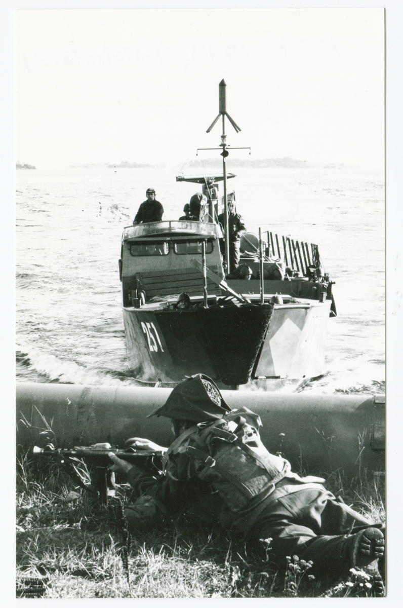 Patrullerande båt vid vattenlinje där militär ligger i skydd
bakom etn rörledning. KFÖ(krigsförbandsövning) 1975,
Vikbolandet.