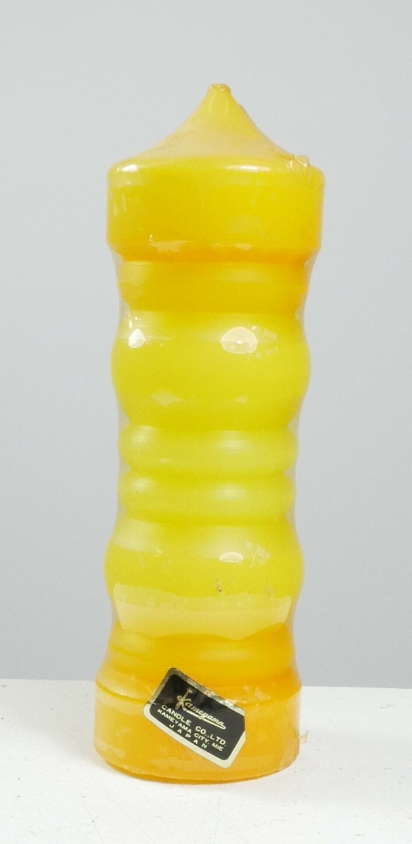 Gul sylinderformet stearinlys pakket inn i plast. Nederst på plasten er det en svart etikett. 

Påskrift, etikett: 
Kameyama // CANDLE CO.. LTD. // KAMEYAMA CITY. MIE // JAPAN 