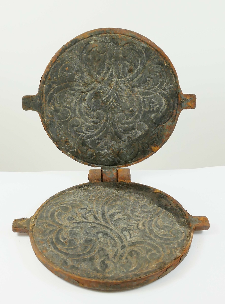 Rundt krumkakejern av metall. Består av to runde jernplater som er henglset sammen bak. Øverst framme på den ene platen er det et håndtak. 