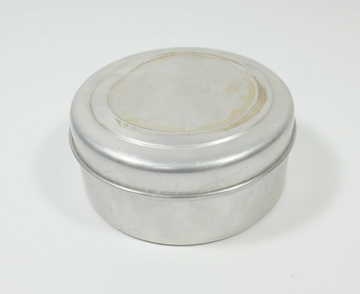 Rund grå beholder av metall med lokk. Inne i beholderen er syltevoks med en rund papirbit med bruksanvisning. 