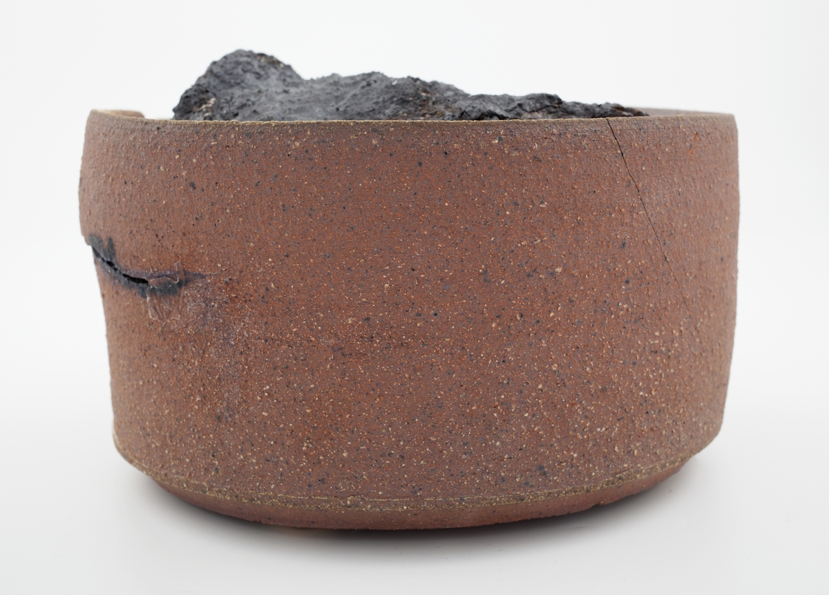 Sylindrisk form i kobberoksidert steingods med en tydelig sprekk nær munningsranden. Inne i beholderen er det en krakellert antrasitt som nesten fyller hele volumet.