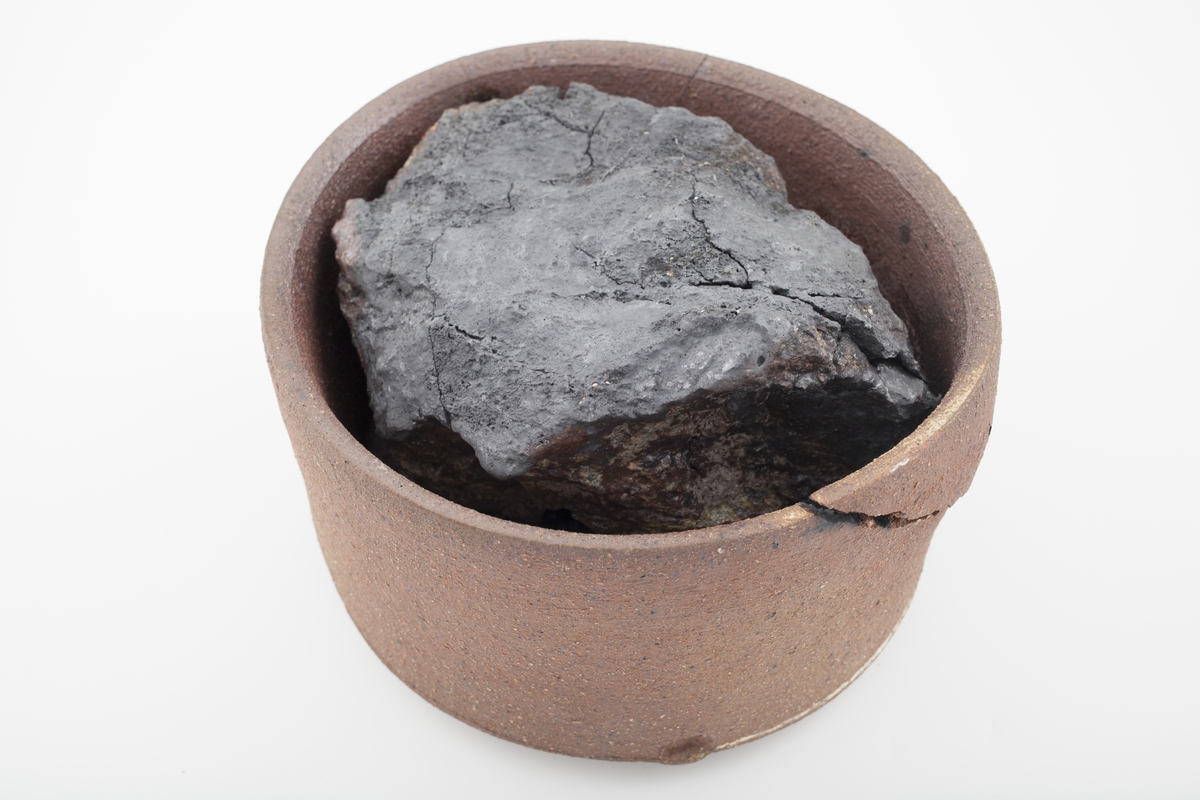 Sylindrisk form i kobberoksidert steingods med en tydelig sprekk nær munningsranden. Inne i beholderen er det en krakellert antrasitt som nesten fyller hele volumet.
