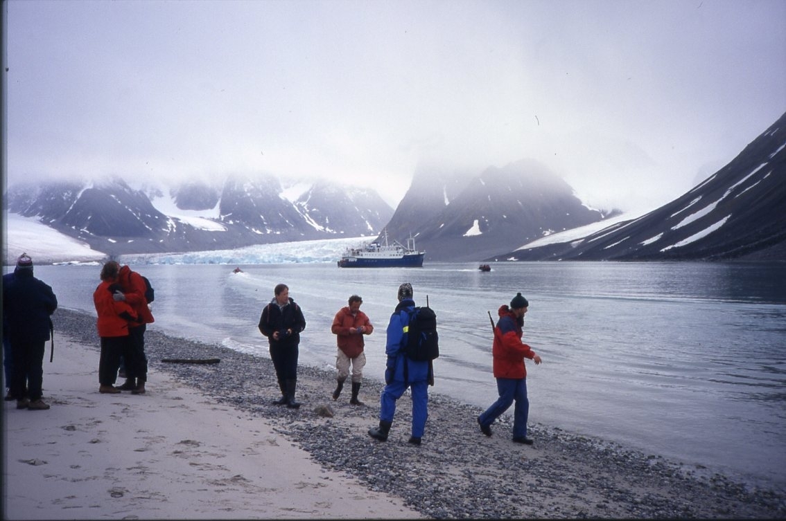 En grupp turister just landsatta på Gravnesodden i Magdalenafjorden. Till höger reseledare och säkerhetsvakt. En zodiac är på väg till och en annan från turistfartyget. Bakom syns Waggonwaybreen. Det är låga moln.