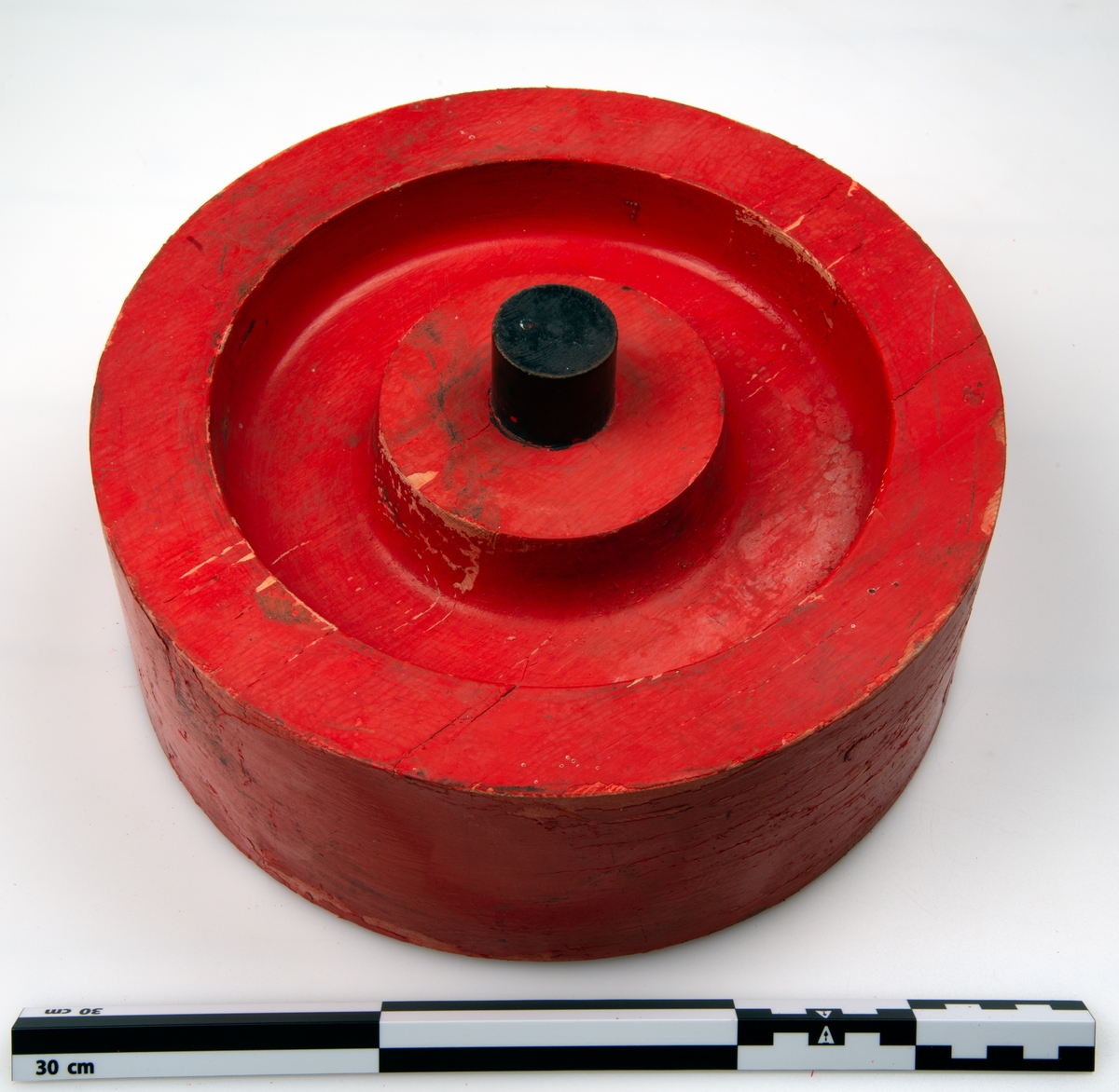 Sirkulær gjenstand, med utsjæringer på en side og utstikkende sylinder på den andre siden. gjenstanden er hovedsalkig malt rød, med detaljer i sort.