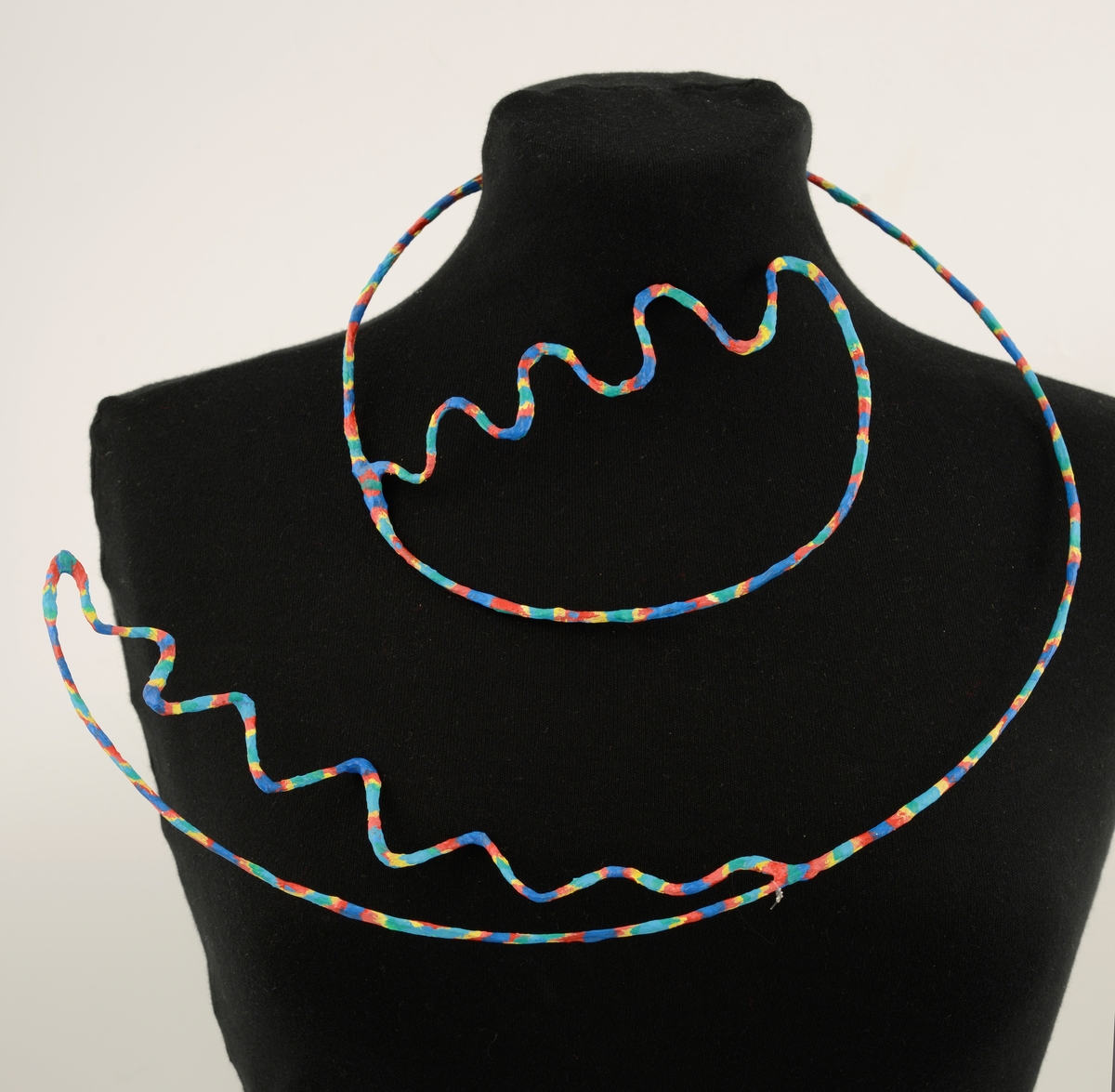 Halssmykke laget av metalltråd som er dekket med papir malt med akrylfarge i forskjellige farger; rød, blå, gul og grønn. Smykket er formet som en åpen spiral som ender i siksakk-former ytterst og innerst.