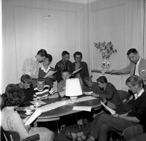 En samling männiksor sitter runt ett bord och sjunger tillsammans. Alla har blivit tilldelade ett texthäfte.