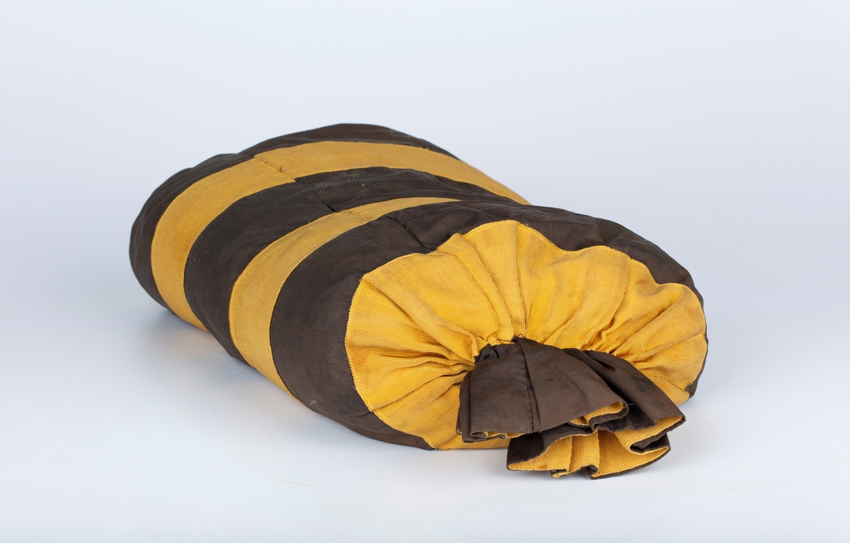 Sylinderpose trekk rundt d pute, laget av striper av brunt stoff og sennepsgule ripsbånd. Endene lukket med rynketråd.