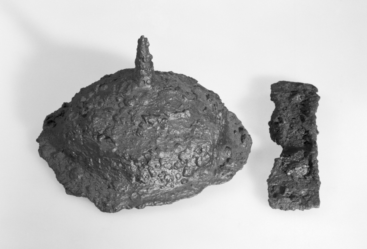 Skjold håndtak av jern, som FETT: Arms in Norway, fig. 62, nå brutt i to deler. Litt tre sees rustet fast. Det er litt skadd i kanten. Lengde 6,5 og 9,4 cm