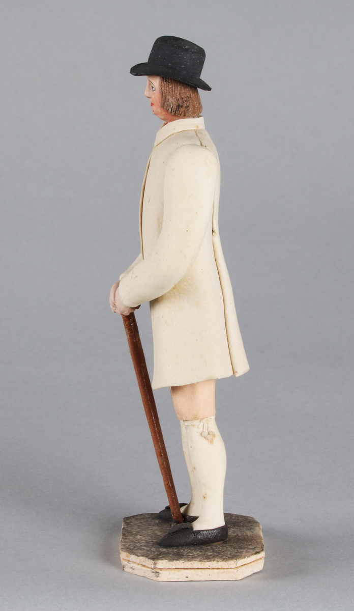 Dragantarbete. Mansfigur.
Allmogedräkt. Stående mansfigur iförd svart hatt med platt kulle och rakt brätte, ej så brett. Rakt kortklippt hår. Halsduk i halsen. Ljus 
ytterrock med långa ärmar, i vars mittparti synes randig väst, röd och svart. Knäkort, ljus, aprikosfärgad byxa, med vita tofsbollar vid knäet. Vita strumpor. Svarta skor. Brun promenadkäpp. Står på grått underlag bestående av åttkantig platta.

Tillverkad av Hilda Boström (1822-1903), sockerbagare, Borås.

Litt: Se Årsbok 1963, sidan 44.

Funktion: Prydnadsfigur