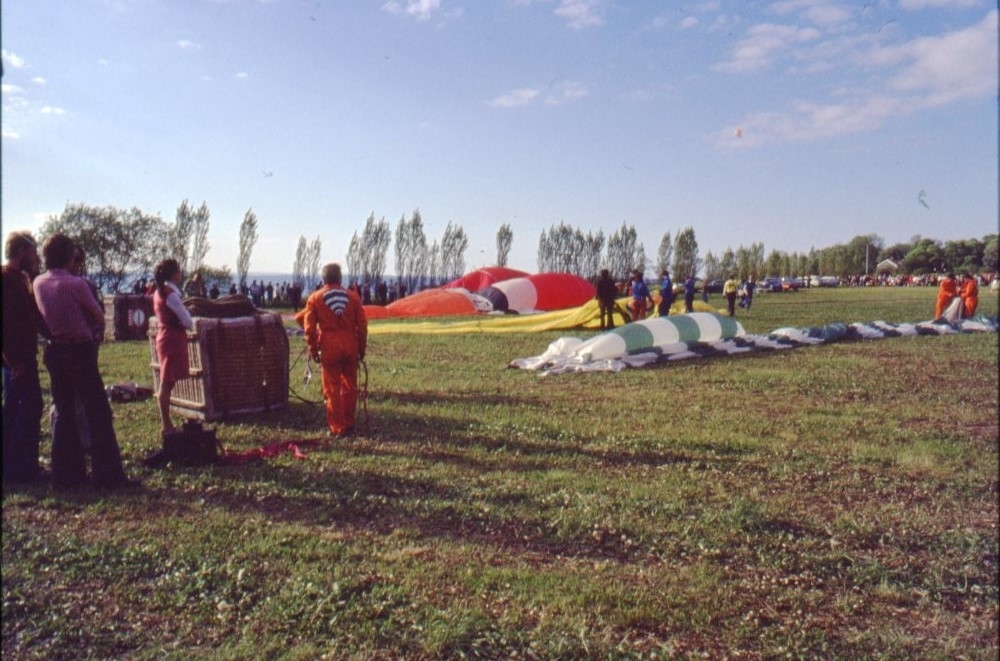 En man och en kvinna står vid en ballongkorg på en gräsplan. Mannen har en orange overall med ballongtryck på ryggen. Bortom dem ligger ett par ballonghöljen utlagda.