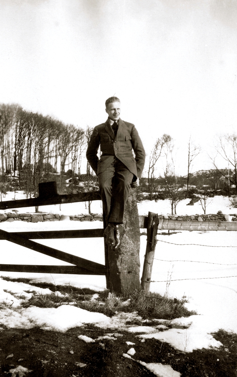 En glad okänd ung man, klädd i kostym, sitter på en grindstolpe vid gärdesgården tillhörande Kållered Stom "Nygård", okänt årtal. I bakgrunden ses ett vinterlandskap.