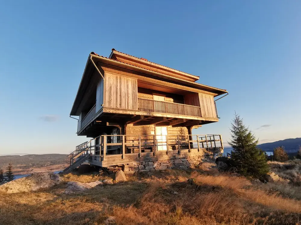 Lysthus på toppen av en kolle i høstfarger. Himmelen bak er blå og sola skinner og speiler seg i vinduet på huset.