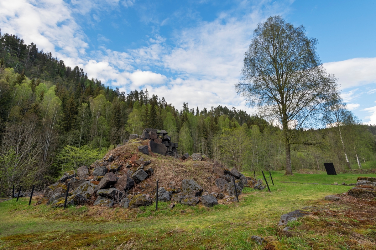Ruinen etter Fritzøe jernverks masovn på Moholt i Siljan. Fotografiet er tatt fra vest. Det rennende elvevatnet som drev blåsebelgene som tilførte den metallurgiske prosessen som foregikk her nok oksygenrik luft til at temperaturen inne i ildstedet ble høy nok, er her skjult bak ruinen, til venstre i bildet.

Masovnteknologien ble innført i Norge tidlig på 1600-tallet. Det dreide seg om digre, høye steinovner som hadde noenlunde runde brennkamre. Når en slik ovn skulle settes i drift, ble den fylt med trekol, som ble antent og fikk stå å gløde 3-4 uker før konstruksjonen var varm nok. Tilstrekkelig høy varme ble oppnådd ved hjelp av oksygentilførsel fra et par blåsebelger som ble drevet ved hjelp av vannkraft fra elva.  Etter at ovnen hadde stått full av glødende trekull et par uker, var murverket varmt nok til at den metallurgiske produksjonsprosessen kunne starte. Det ble gjort ved «påsettinga» av trekol og malm, lag for lag, fra «krona», overkanten på masovnmuren. Slik kunne masovnen holdes i kontinuerlig drift gjennom et par år før det ble nødvendig å avslutte fyringa og skifte ut steinene i de mest varmeeksponerte delene av brennkammeret. Dette forutsatte naturligvis at verket hadde gode lagere av malm og trekol. Mens prosessen pågikk ble det stadig tappet råjern gjennom et hull - «stellet» - i den nedre delen av ildstedet. Dette jernet kunne enten brukes i støpejernsprodukter umiddelbart, eller det kunne bearbeides videre til smidigere smijern i en «hammer».  På Moholt ble det bygd et hammerverk tidlig i 1730-åra. Hit transpporterte man forholdsvis små volumer av råjern fra Fritzøe-verkets hovedanlegg i Larvik med ferger over innsjøen Farris, i stedet for å frakte store volumer av trekull den motsatte vegen. Under napoleonskrigene tidlig på 1800-tallet økte etterspørselen etter jern. I denne perioden hadde det vist seg vanskelig å skaffe nok trekull til masovnen i Barkevik i Brunlanes. Derfor bestemte ledelsen i Fritzøe-verket at noe av råjernproduksjonen skulle flyttes til Moholt, der man hadde bedre tilgang på trekull fra skogene i Siljan. Dette var bakgrunnen for at man bygde den masovnen vi ser på dette fotografiet i perioden 1809-1817. Årsaken til at byggeprosessen tok så lang tid var at de varmeresistente steinene som skulle være i området rundt stellet måtte importeres, noe som var vanskelig i en periode med krig og blokader av skipsfarten. Masovnen på Moholt var i drift i en 50-årsperiode, fra 1817 til 1867. Den var omgitt av ei smeltehytte, en bordkledd bindingsverkskonstruksjon, som vi ser noen av fundamentene til i forgrunnen på dette bildet. Dette kulturminnet er tilgjengeliggjort for interesserte med informasjonsskilt og tilstøtende rasteplass. Klatring i ovnsruien er ikke ønskelig. Den er derfor skjermet ved hjelp av et enkelt kjettingstengsel.   

Litt mer historisk informasjon om den metallurgiske virksomheten på Moholt finnes under fanen «Opplysninger».