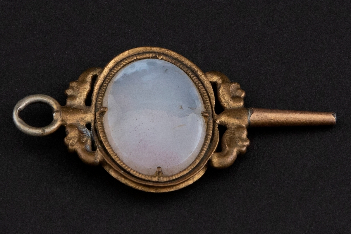 Urnøkkel av forgylt messing. I midten er en oval gråhvit agat i riflet ramme. Den er omkranset av støpte ornamenter, to oppe og to nede.