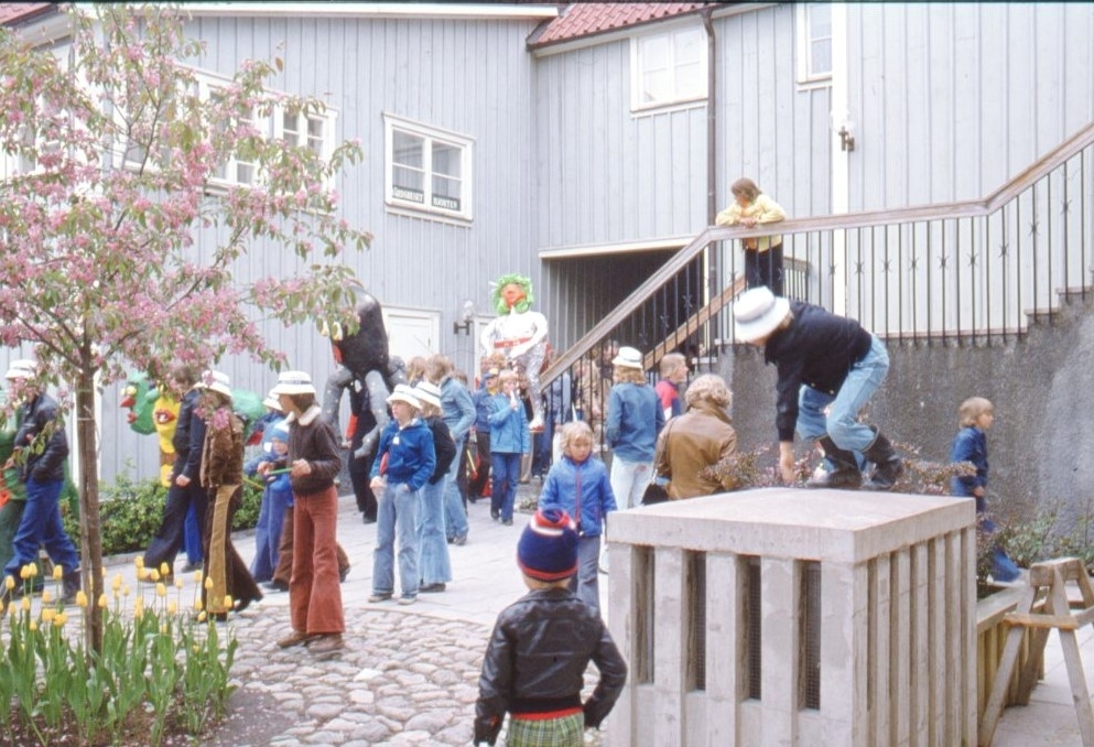 Barn med stora papier-mache figurer på innergården till kv Hjorten söder om Gränna torg. En person går runt i gammaldags polisuniform. Troligen i samband med arrangemanget "Barnens stad".