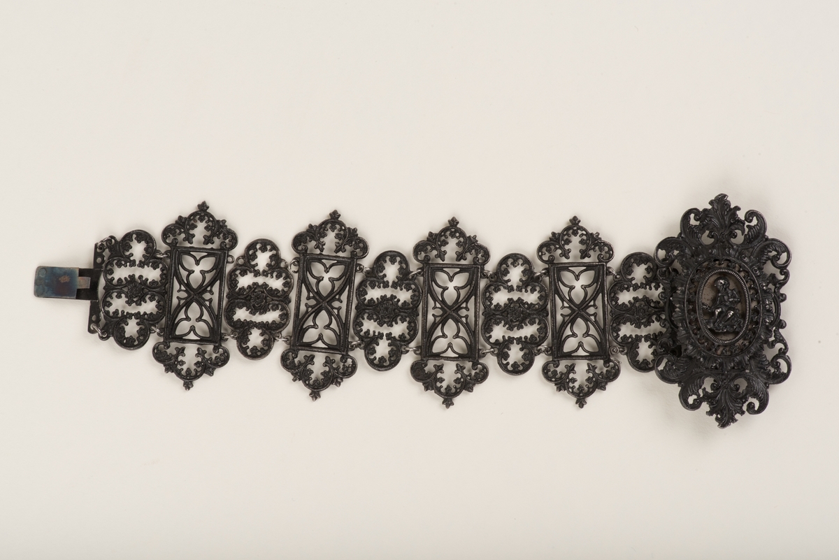 Svart armband av tio ledade länkar av gjutjärn.
Länkarna är utformade som gotiska trepass dekorerade med växtornament i rokokostil. Länken som döljer låset är större än de övriga och dekorerad med ett sittande figur som framhävs genom en förnicklad plåt under.
