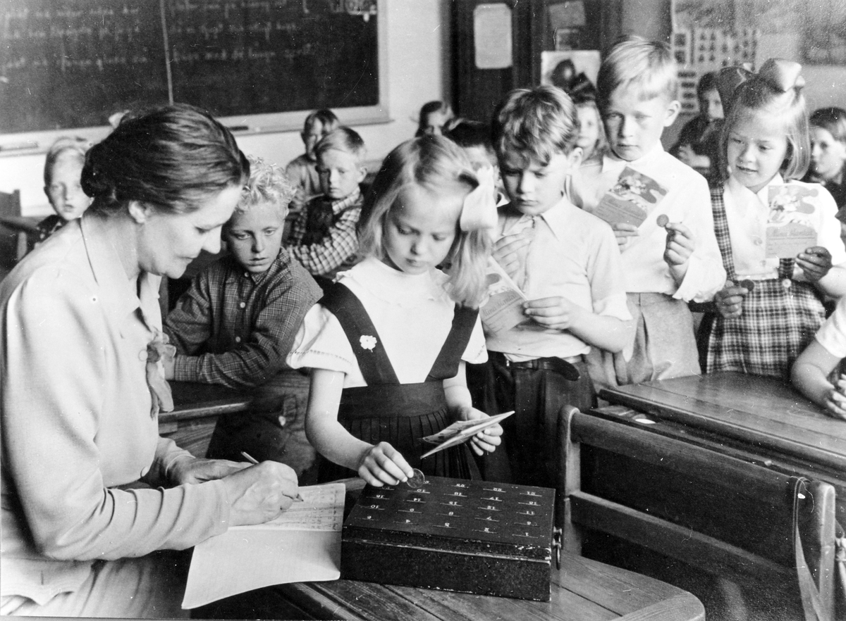 Skolsparande på Centralskolan. Lärarinna Karin Johansson och elever i ett klassrum, några barn står i kö med mynt till sparbössa.
