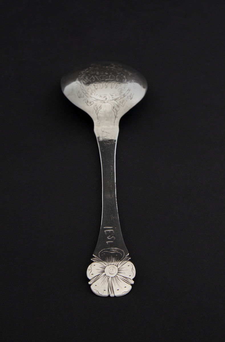 Barokkskje i forgylt sølv med flatt skaft og ovalt skjeblad. Skaftet avsluttes med gravert fembladet rosett. Baksiden av skjebladet er dekorert med en kartusj formet som en krans.