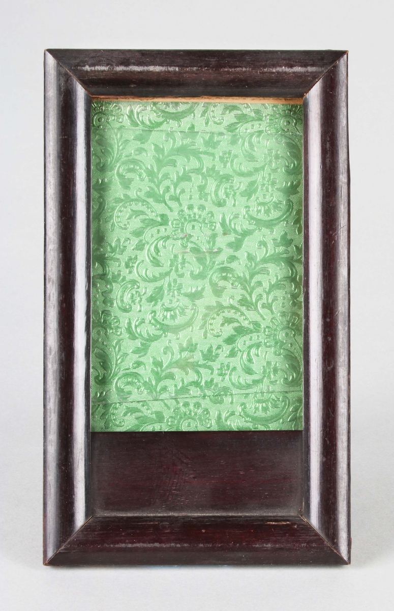 Fotografiram. Rektangulär, med glas inom brunmålad träram. Bakstycke av papp med fotstöd. På framsidan, pålimmat, grönt, reliefpressat papper med bladmönster.