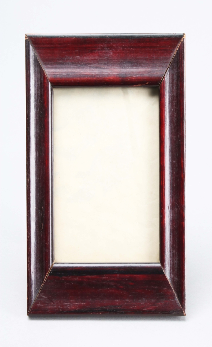 Fotografiram. Rektangulär, med glas inom mahognybetsad träram. Bakstycke av papp med fotstöd. På framsidan, bakom glaset, pålimmat, beigefärgat papper.
