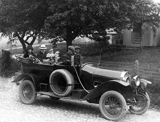 En bil (cabriolet) står på Brahegatan i Gränna, vid torgets södra uppfart, riktning söderut. I bilen sitter två män fram samt en kvinna och en man i baksätet, med blommor. Mannen bakom ratten är troligen doktor Eurén från Jönköping.