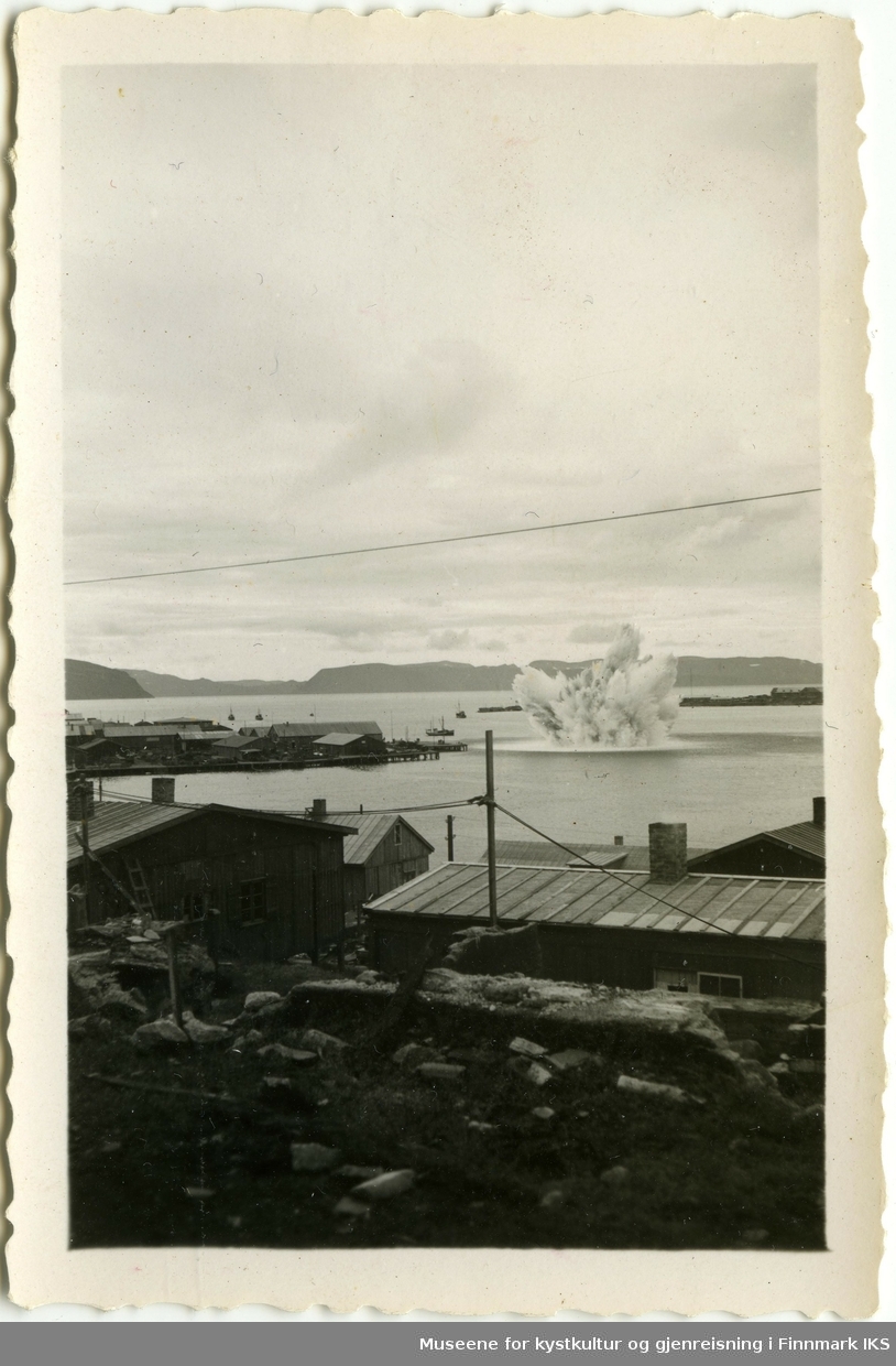 En sjømine detonerer i havnebassenget i Hammerfest. Til venstre og i forgrunn ser man brakkebebyggelse. Ti høyre i bakgrunn ser man Fuglenesodden, til venstre Håja-fjellet i Sørøysundet.