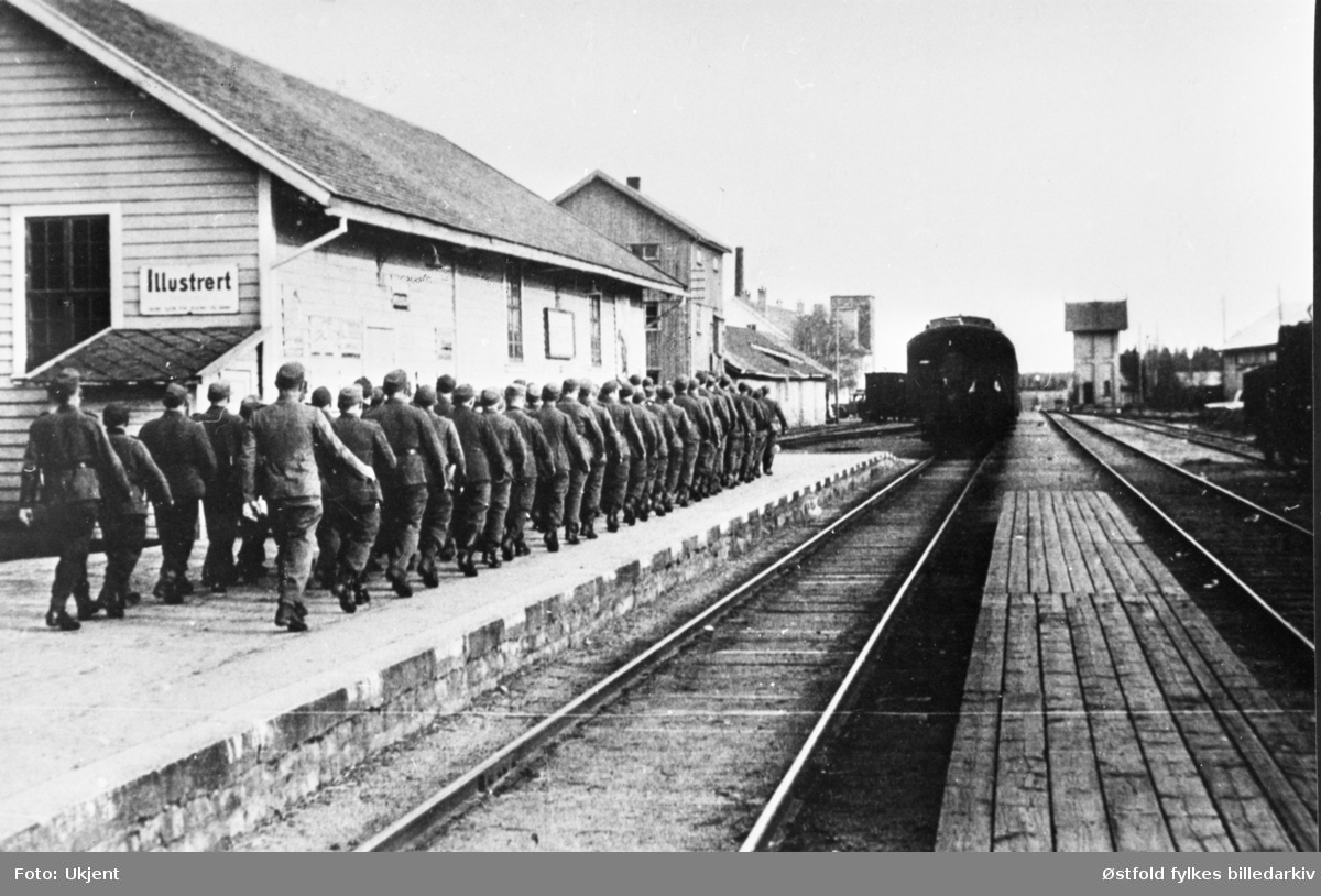 Tyske soldater masjerende inn på Rakkestad jernbanestasjon mai 1945. De reiser fra bygda. Reklameskilt for illustrert.