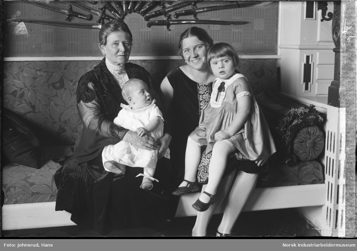 To kvinner sittende med hvert sitt barn på fanget i sofa. Knivdekorasjon på veggen i bakgrunnen. 
Fotografi bestilt av Konsul Coward.