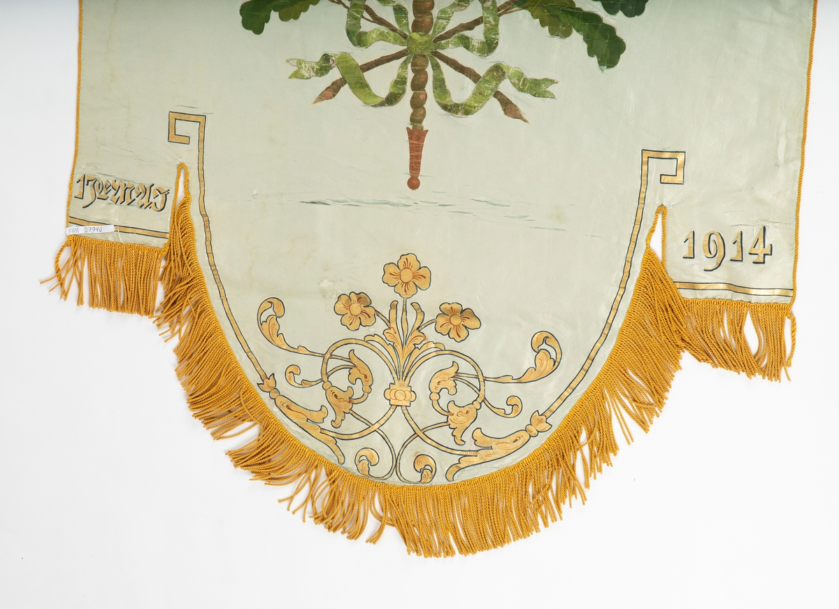 Skolefane. Silke. Hvitt stoff. 
Håndmalt dekor: Grønn eikeløvkrans rundt fakkel. Drøbak Folkeskole. 1914.
Stilisert rankemønster i bue nederst. Gyldengule silkefrynser. 
Fanen tredd inn på tverrstang av tre.