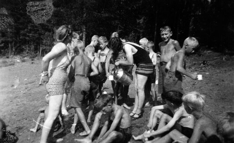 Gutter fra Borgen feriekoloni på badeplassen ved Fangberget i Mjøsa, 1940. Saft er medbrakt på melkespann og serveres til barna. De voksne som er med på turen er bestyrerinnen, kokka og stuepiken. Foto: Anno Domkirkeodden. (Foto/Photo)