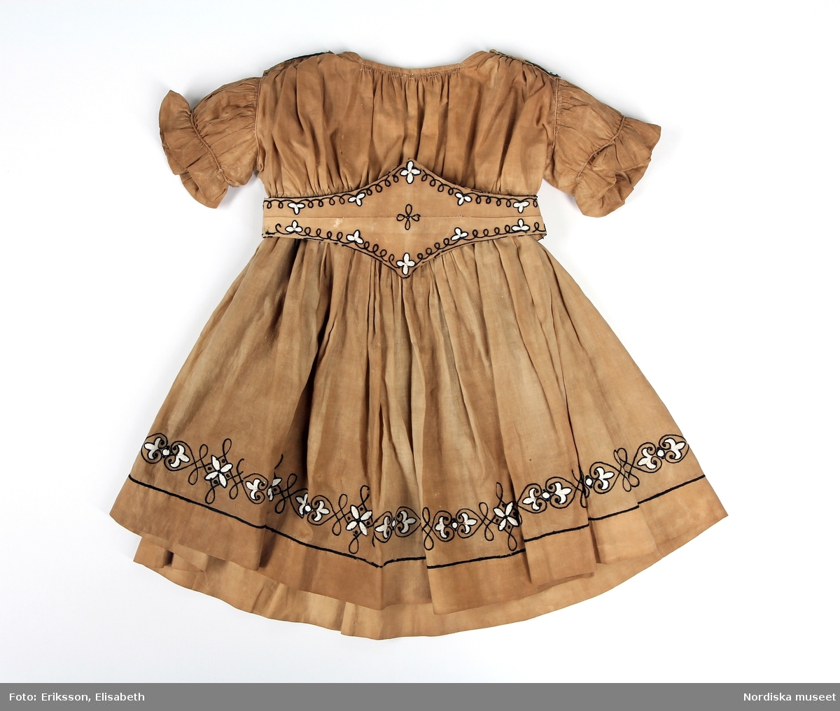 Huvudliggare:
"Barnklädning, bestående av: liv, kjol och skärp av gult bomullstyg med svarta och vita broderier. Början av 1860-talet:"