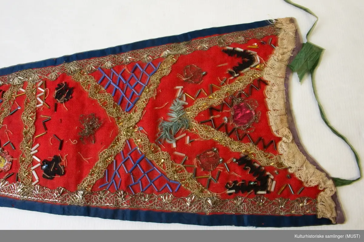 Dåpssmekke av rødt tøy kantet med blå silke, gull- og sølvborder og perlepynt. Grønne og røde pyntebånd. For av blåmønstret tøy.