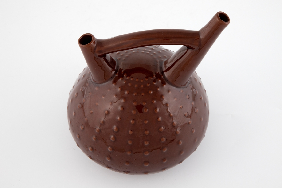 Vase i leirgods, dekket av brunrød glasur. Flattrykket kuleformet korpus med radiære "nupper", hvis utforming alluderer til en kråkebolle. Fra øvre del av korpus reiser det seg to tuter, som er forbundet med hverandre via en hank.