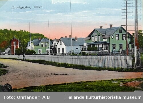 Från Järnvägsgatan i Ullared. Till höger närmast kameran syns en fällbar bom för korsningen Allmäna vägen genom samhället och "Varbergsbanan" - Varberg-Ätrans Järnväg. Gröna byggnaden bredvid uppfördes som hotell vid den tidpunkt det bestämdes att Ullared skulle bli järnvägsknut. Huset revs ca 1980. Det vita huset är apoteksbyggnaden och den gröna byggnaden därnäst är J A Johanssons lanthandel. Näst sista byggnaden i raden, benämt vattentornet, tjänade som pumpstation för ångloken på Varbergsbanan. Vatten togs från Musasjön. Sista byggnaden användes som banmästarebostad.