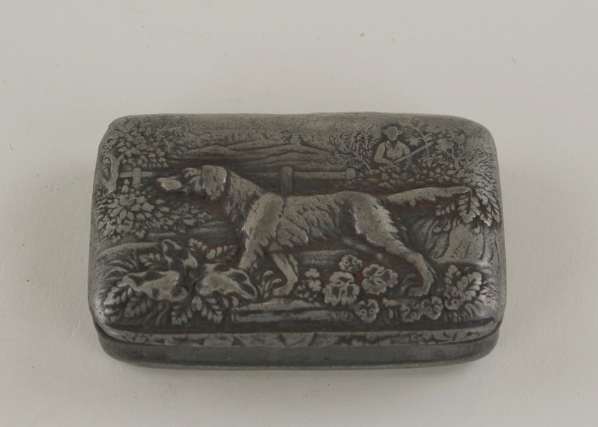 Rektangulär snusdosa av plåt. 
På locket finns ett motiv i relief av av en hund som står i ett landskap. I bakgrunden en man med gevär.
Dosans sidor är något insvängda.