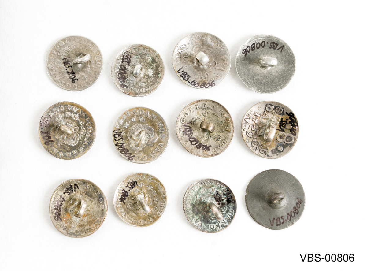 Samling av 21 sølvknapper, i forskjellige størrelser som er omlagd av shilling mynter.
Avrundete i form, i det indre konvekse området, har de en liten sveiset ring å sy til plagget